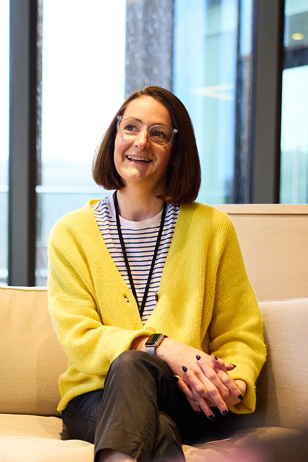Zenith colleague sat on sofa with mid-length brown hair and glasses sat smiling with a bright yellow jumper on with her hands crossed over her knees